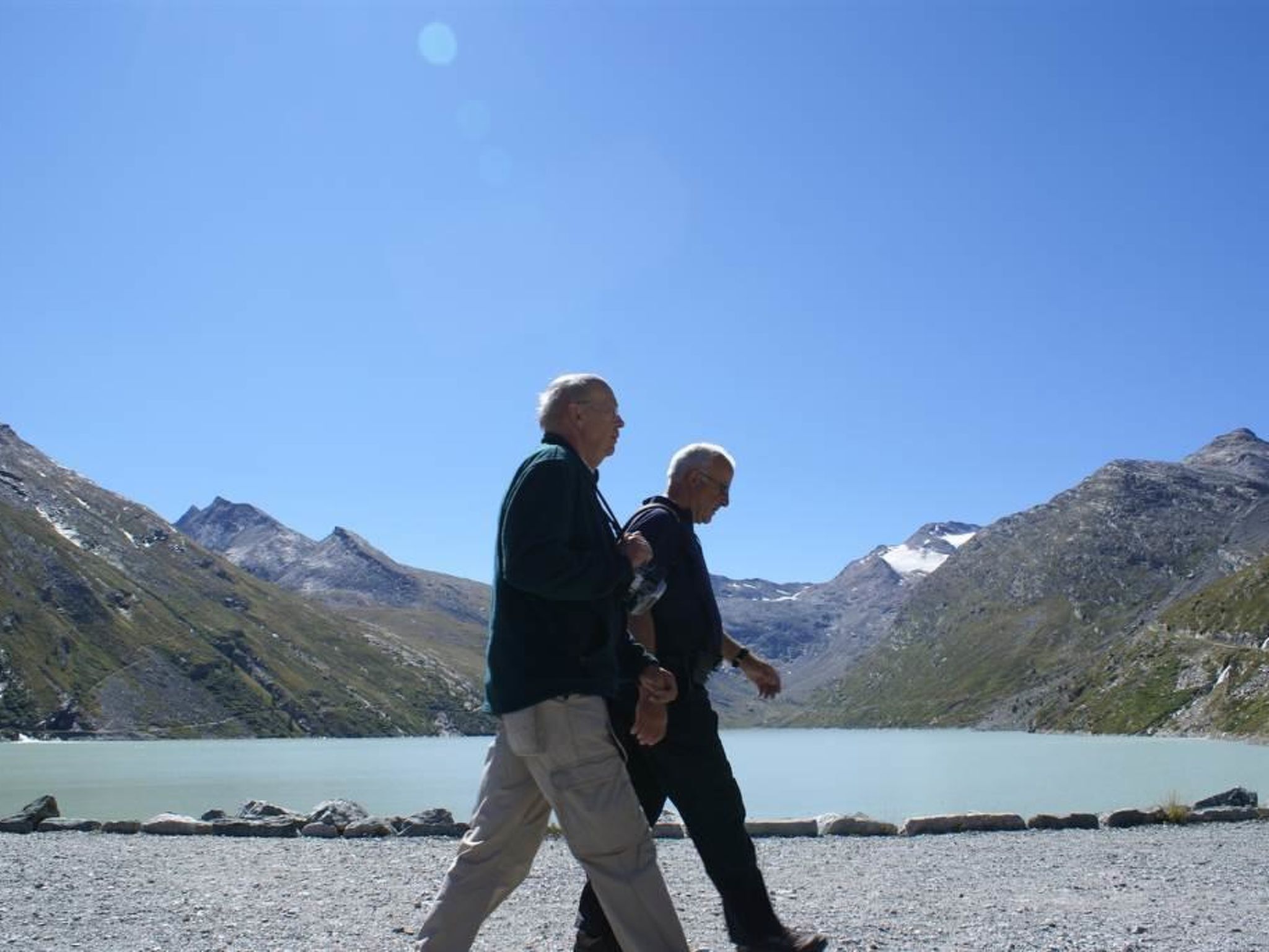 Two people walking on a gravel path with a mountainous backdrop and a lake.