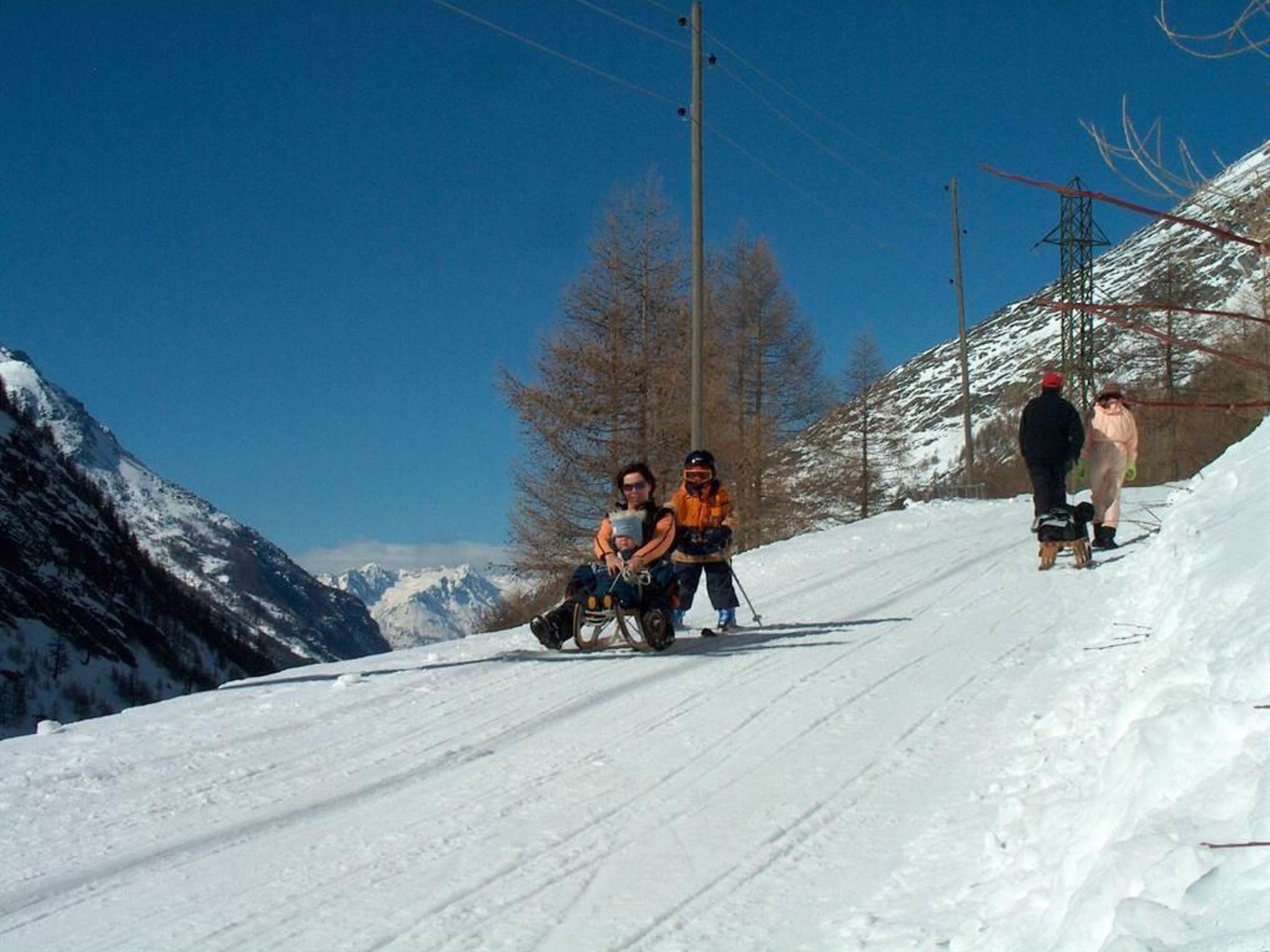 A snowy mountain slope with a person in a wheelchair and another person in an orange jacket.