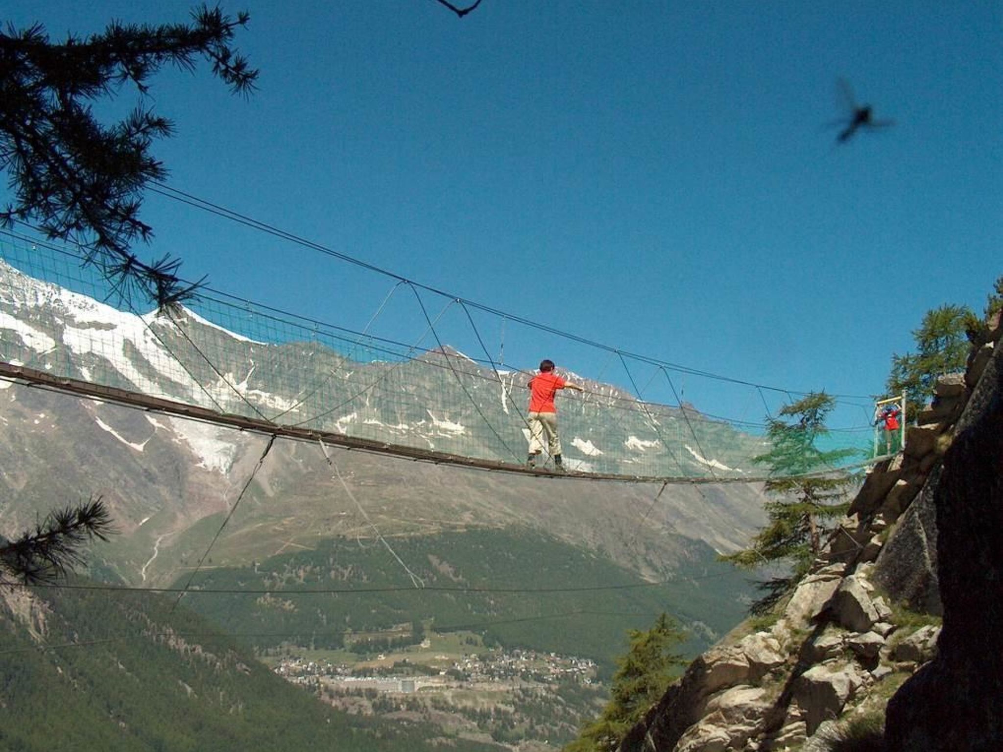 A person walking on a suspended bridge with a mountainous landscape in the background.