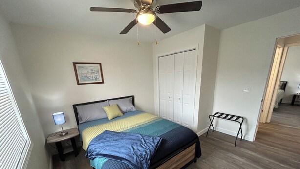 A bedroom with a bed, a ceiling fan, a nightstand, a lamp, a door, and a small table with a chair. - "The Hive" Cottage #1 (Van Buren)