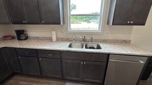 A kitchen with dark cabinets, a granite countertop, a stainless steel sink, and a dishwasher. - "The Hive" Cottage #1 (Van Buren)