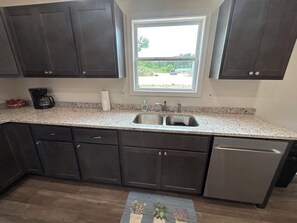 A kitchen with dark cabinets, a granite countertop, a stainless steel sink, and a dishwasher. - "The Hive" Cottage #1 (Van Buren)