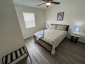 A bedroom with a bed, a ceiling fan, a window with blinds, a bedside table with a lamp, and a chair. - "The Hive" Cottage #1 (Van Buren)