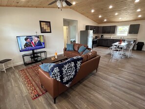 A modern living room with a brown leather sofa, a television displaying a news broadcast, and a kitchen area with dark cabinets and a window. - "The Hive" Cottage #1 (Van Buren)