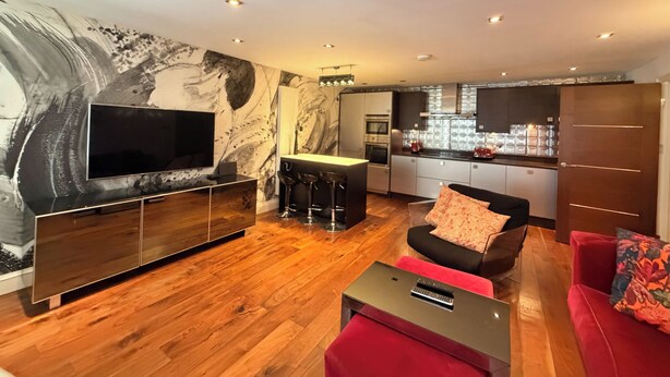 A modern living room with a flat-screen TV, a red sofa, a coffee table, and a kitchen area with a bar. - Artsy and Spacious 2BDR Flat in Central London (Greater London)