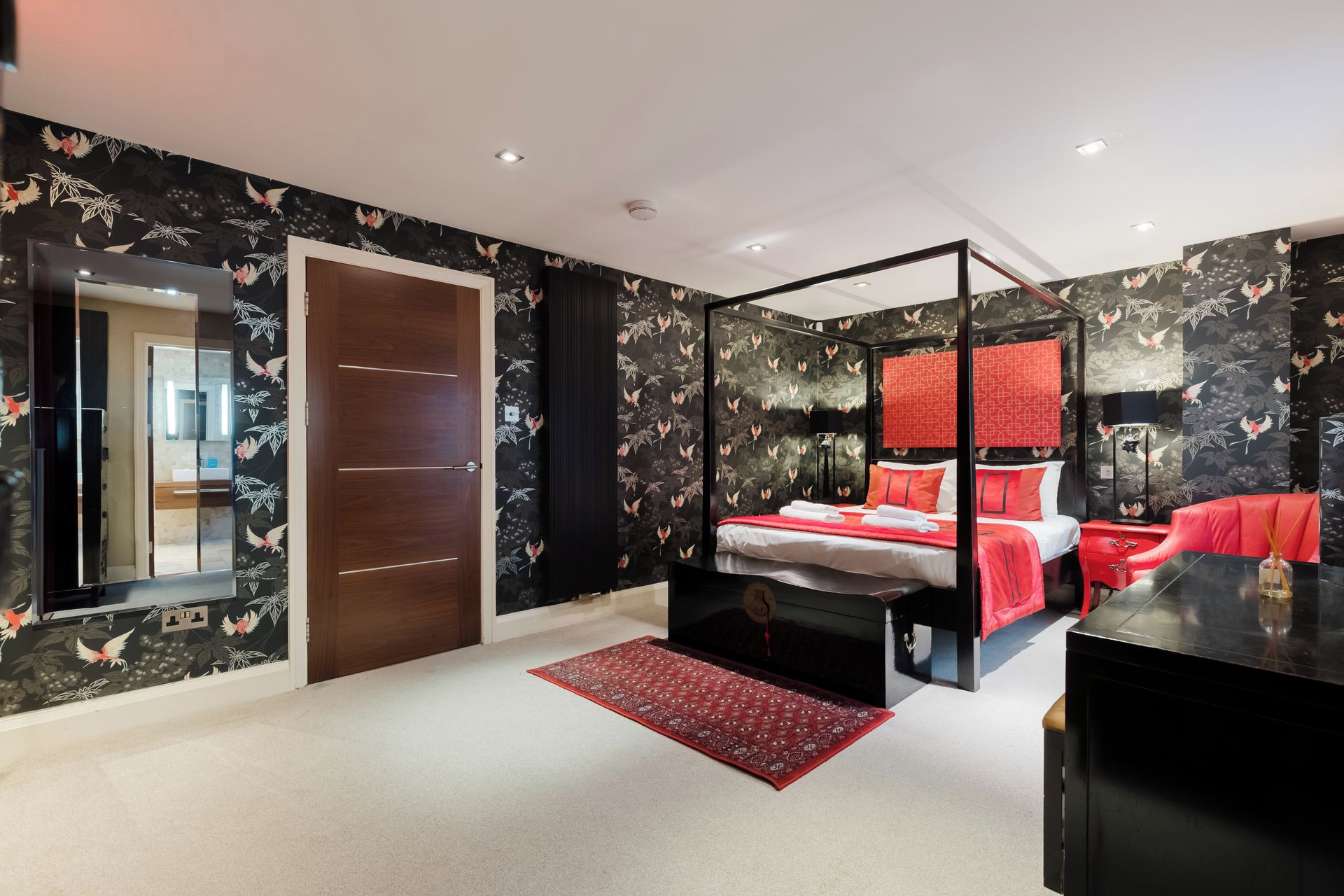 A modern bedroom with a four-poster bed, a red accent wall, and a black and white floral wallpaper.