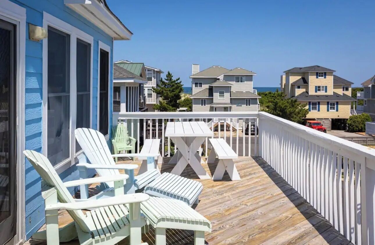 A wooden deck with Adirondack chairs and a picnic table, overlooking a residential area with houses and a clear sky.