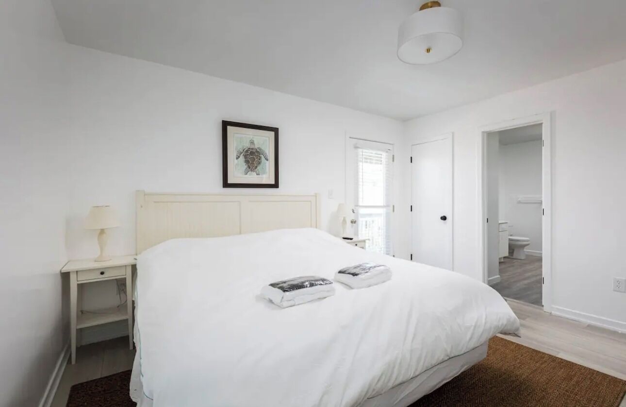 A bedroom with a large bed, a bedside table with a lamp, a framed picture on the wall, and a door leading to a bathroom.