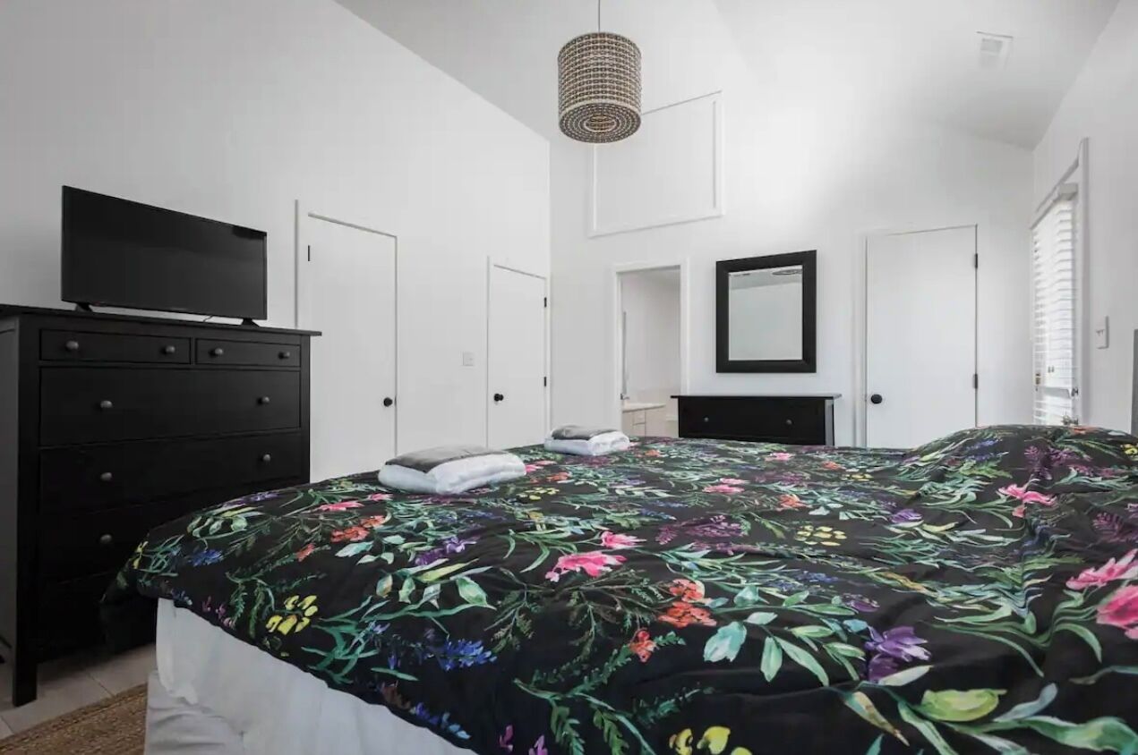 Un dormitorio con colcha estampada, cómoda negra, televisor encima y lámpara colgante.