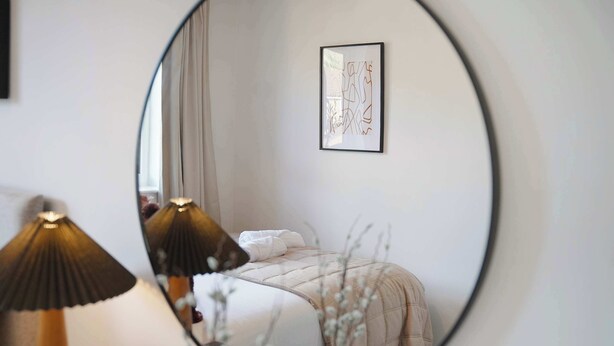 Ein Schlafzimmer mit einem Bett, einer Nachttischlampe, einem gerahmten Bild an der Wand und einem runden Spiegel, der den Raum reflektiert.