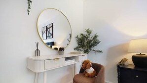 Ein Zimmer mit einem weißen Schreibtisch, einem runden Spiegel, einer kleinen Topfpflanze, einer Lampe und einem braunen Stuhl.