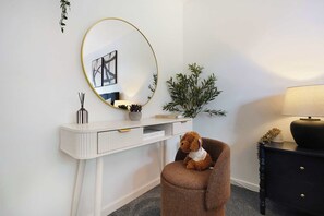 Ein Zimmer mit einem weißen Schreibtisch, einem runden Spiegel, einer kleinen Topfpflanze, einer Lampe und einem braunen Stuhl.