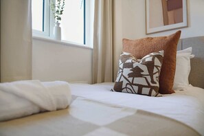 Cama bem arrumada com lençóis brancos e duas almofadas decorativas, uma com padrão geométrico e outra lisa.