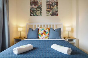 Quarto de hotel com cama, duas mesas de cabeceira, duas luminárias, cabeceira branca e dois quadros na parede.