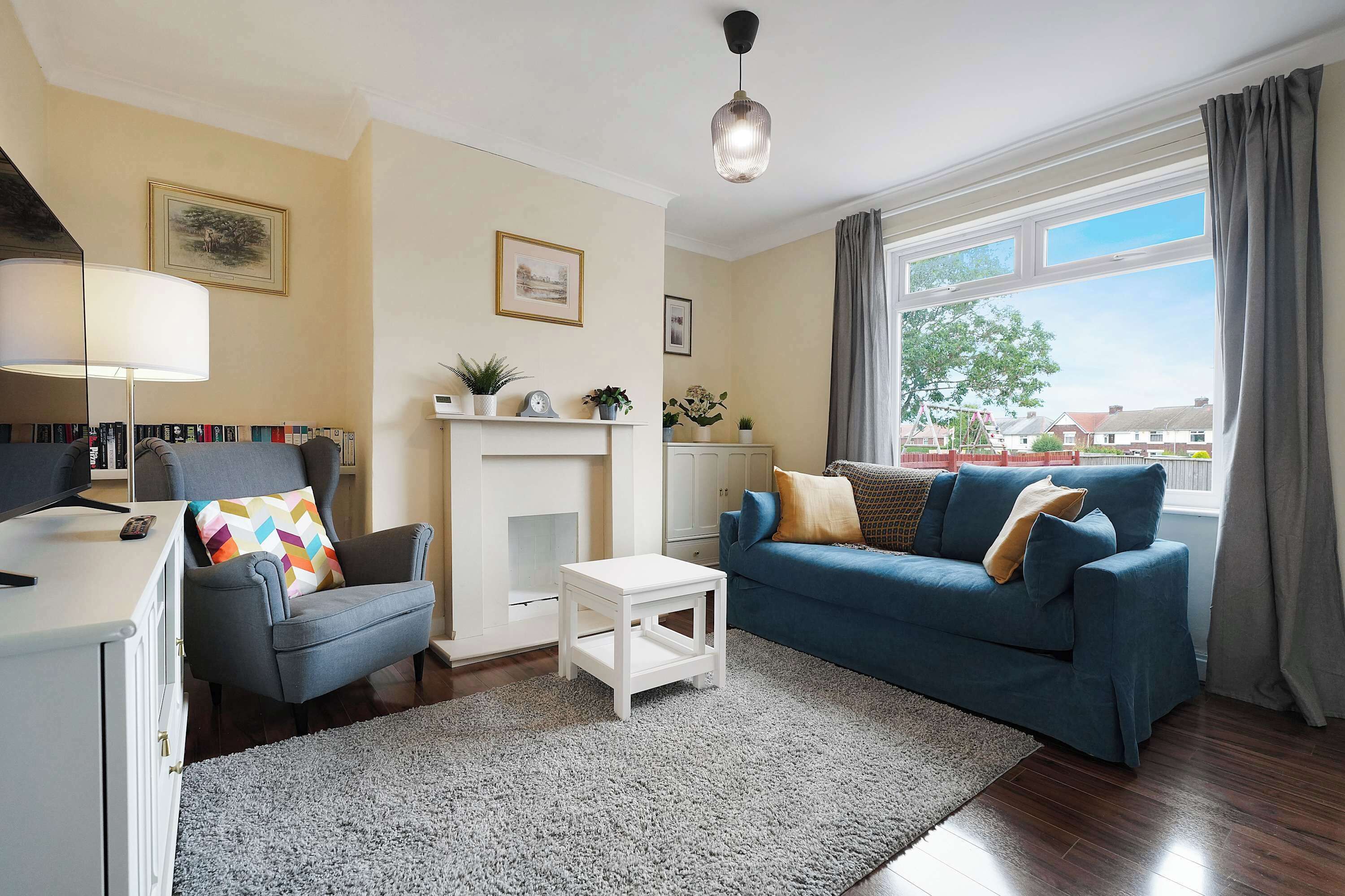 A living room with a fireplace, a sofa, a chair, a small table, and a lamp. There are bookshelves and a window with curtains.