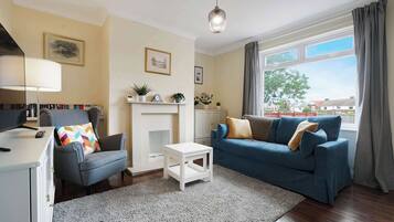 A living room with a fireplace, a sofa, a chair, a small table, and a lamp. There are bookshelves and a window with curtains.