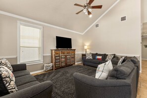 Ein Wohnzimmer mit Deckenventilator, Flachbildfernseher, Holzschrank und grauem Sofagarnitur.