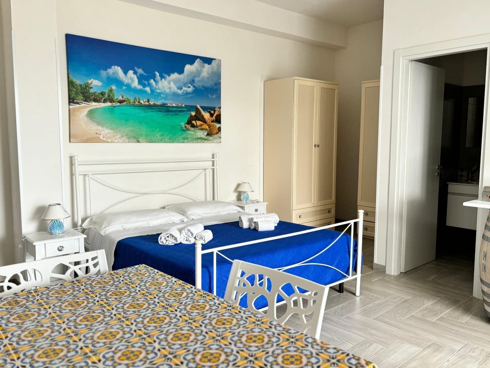 Une chambre d’hôtel avec un lit, un bureau, une armoire et un tableau représentant une scène de plage.