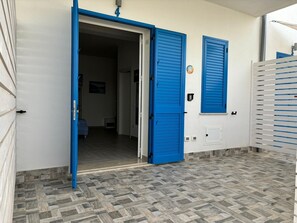 Een open deur die leidt naar een hal met een patroon in de vloer, een blauwe luik en een witte muur met een klein plaatje.