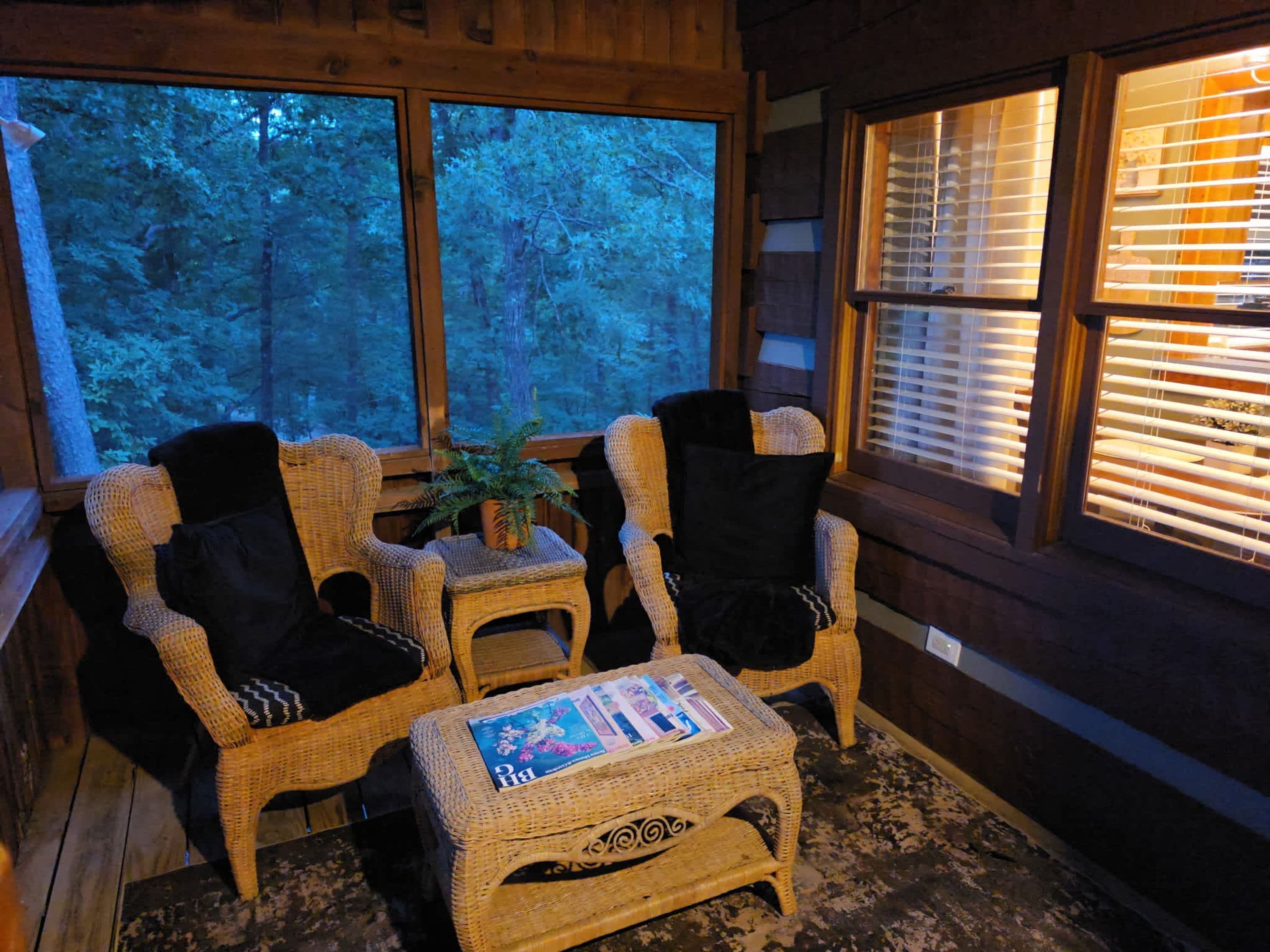 Una habitación acogedora con muebles de mimbre, una mesita con una revista y vistas a árboles a través de amplias ventanas.
