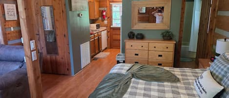 A cozy cabin interior with a bed, a dresser, a mirror, a kitchen area, and a bathroom.