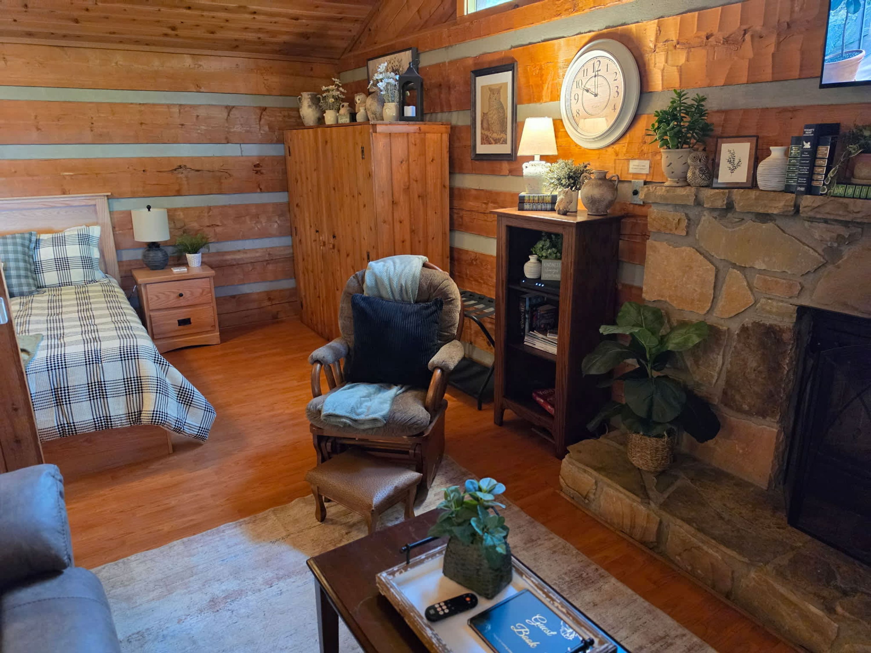 A cozy cabin interior with a fireplace, wooden walls, a comfortable armchair, a bed with plaid bedding, and a bookshelf.