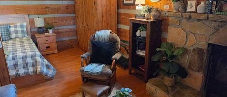 A cozy cabin interior with a fireplace, wooden walls, a comfortable armchair, a bed with plaid bedding, and a bookshelf.