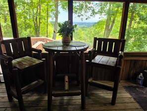 Una terraza de madera con una mesa redonda y dos sillas, una planta en maceta sobre la mesa, y vistas al verdor a través de grandes ventanales.