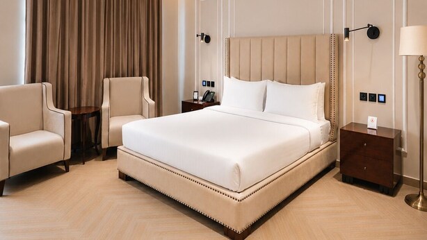Habitación de hotel con una cama grande, dos sillones, una mesita de noche, una lámpara y una cortina.