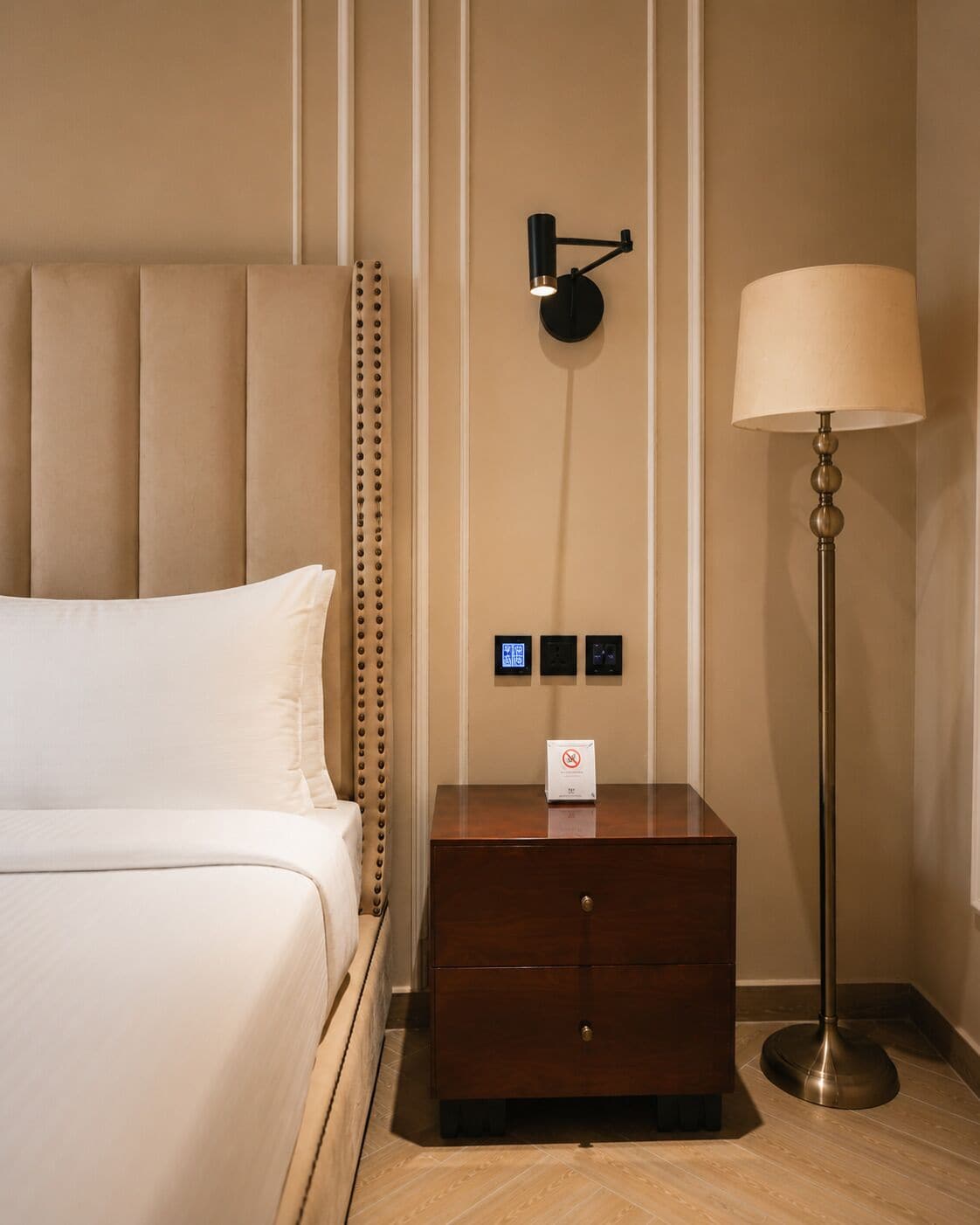 Habitación de hotel con cama, mesita de noche con lámpara y panel de control en la pared.