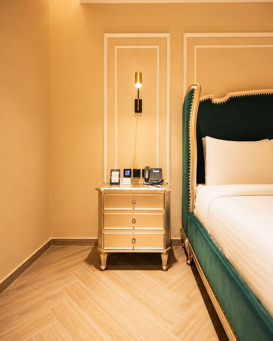 Quarto de hotel com uma cama, mesa de cabeceira com telefone e luminária fixada na parede.
