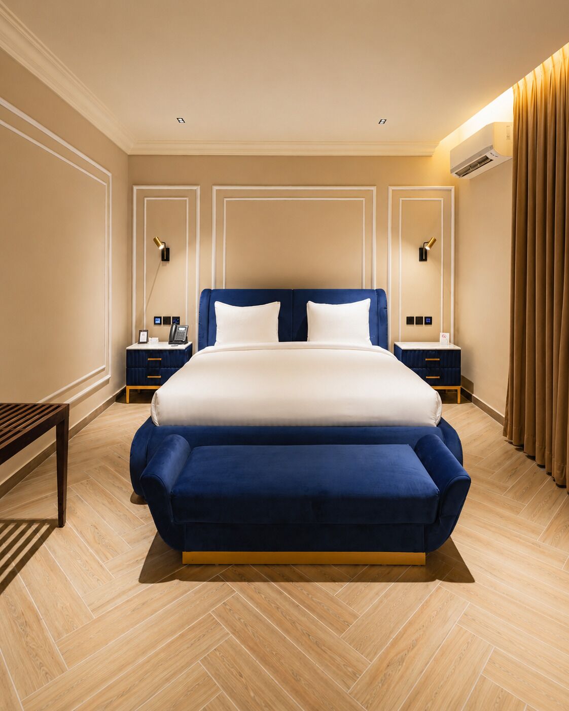 Ein großes Bett mit blauem Kopfteil und weißer Bettwäsche, an beiden Seiten zwei Nachttische mit Lampen, in einem Zimmer mit Holzboden und beigen Wänden.
