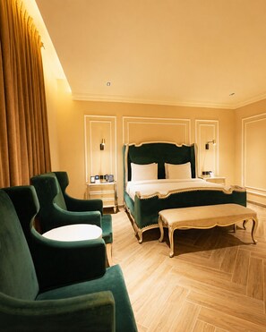 A hotel room with a large bed, two bedside tables, a bench, and green upholstered chairs.
