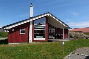 Ein rotes, eingeschossiges Haus mit einem großen Fenster, einem Schornstein und einer kleinen Veranda mit Gartenmöbeln.