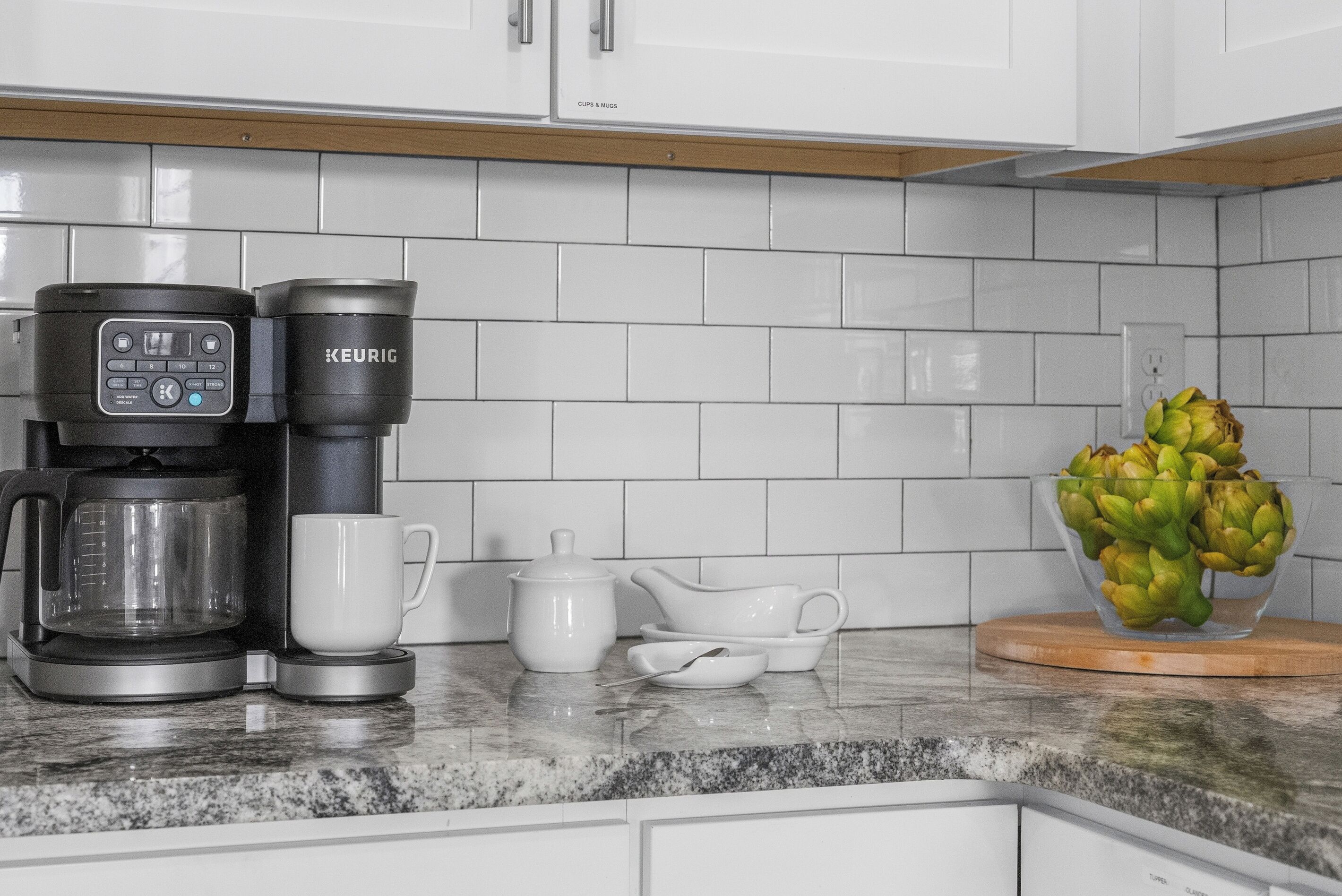 Een keuken met een koffiezetapparaat, een mok, een theepot, een kom en een vaas met artisjokken.