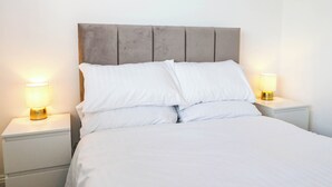 Un llit amb roba de llit blanca, un panell de capçal tapissat de gris i dues làmpades de nit sobre taulells blancs.