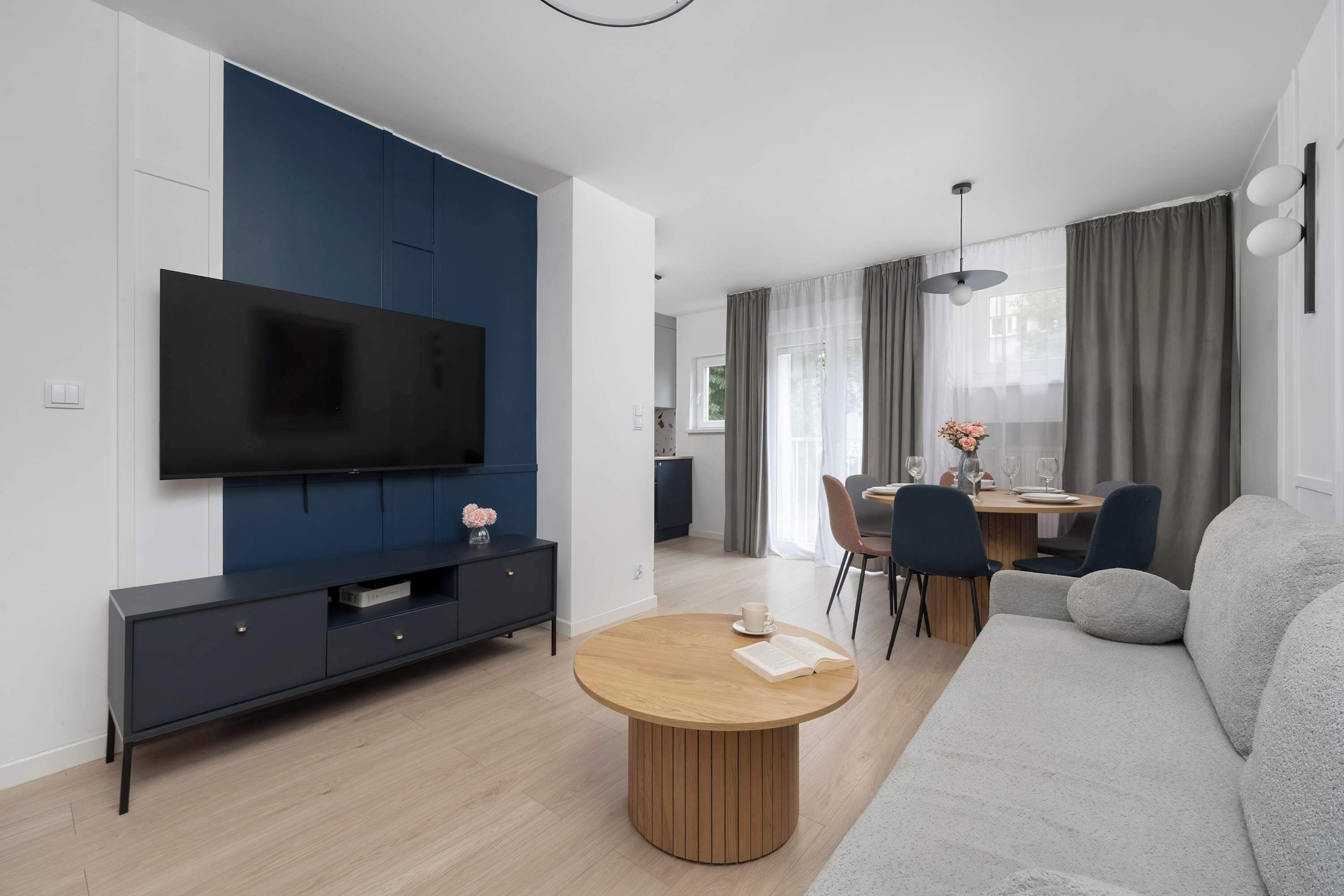 Un salon moderne doté d’un téléviseur à écran plat, d’un mur d’accent bleu foncé, d’une table basse ronde en bois, d’un canapé gris et d’un coin repas avec une table et des chaises.