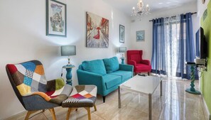 Ein Wohnzimmer mit einem blaugrünen Sofa, einem roten Sessel, einem farbenfrohen Sessel, einem Glastisch und einer Pendelleuchte.