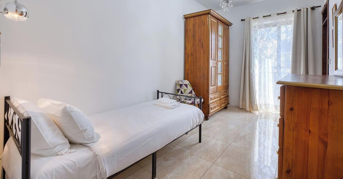 Ein Einzelbett mit weißer Bettwäsche, ein Holzschrank, ein kleiner Tisch mit Lampe und ein Fenster mit Vorhängen.