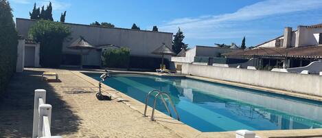A swimming pool with a white fence in the foreground, surrounded by buildings and trees under a clear sky.