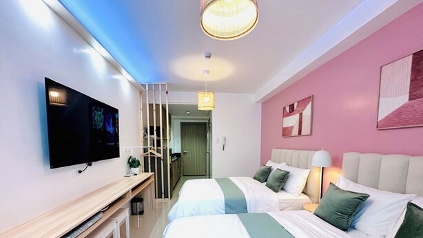 Una habitación de hotel moderna con televisión, escritorio, dos camas, lámpara colgante y pared rosa.