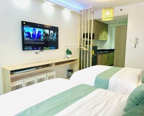 A hotel room with a TV mounted on the wall, a bed with green and white bedding, a small plant on a shelf, and a kitchen area visible through a partition. - Chic 2-bed Family Suite Shore Residences  Pasay City MOA near airport/NAIA/Okada (Pasay City)