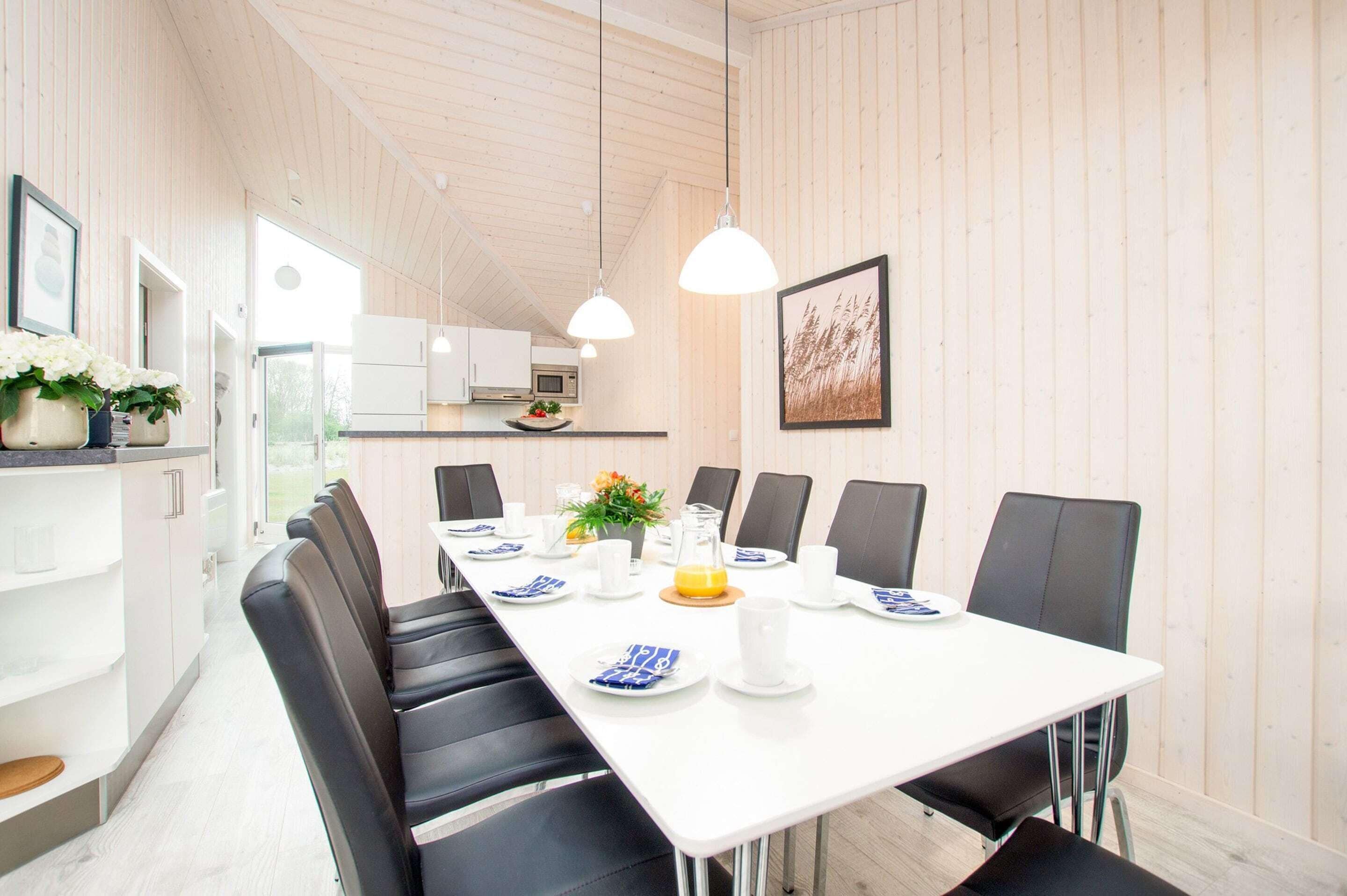 Un espace repas moderne avec une longue table, des chaises noires, de l’assiette blanche et une cuisine en arrière-plan.
