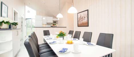 A modern dining area with a long table, black chairs, white plates, and a kitchen in the background.