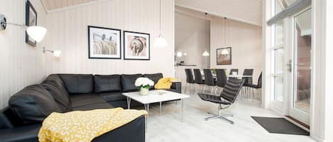 Un moderno salón con un sofá negro, una mesa de centro blanca y una manta amarilla. Hay cuadros enmarcados en la pared y una zona de comedor con sillas y una mesa.