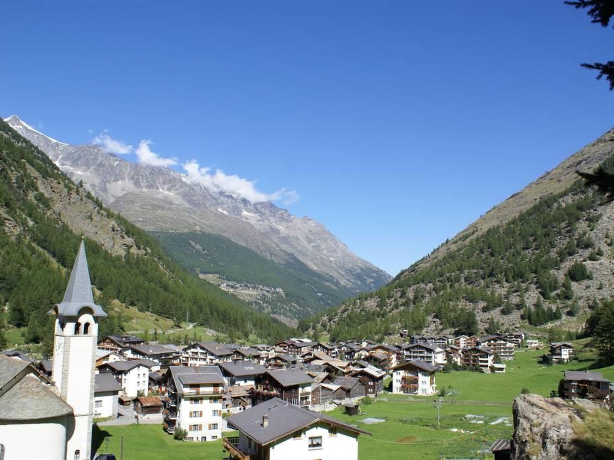 Ein Bergdorf mit traditionellen Chalets, einem Kirchturm und einem Tal mit Bergen im Hintergrund.