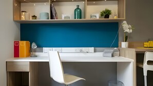 Ett modernt skrivbord med en vit stol, en lampa och en dator. Ovanför skrivbordet finns hyllor med diverse föremål, och en vägg är accentfärgad i blått.