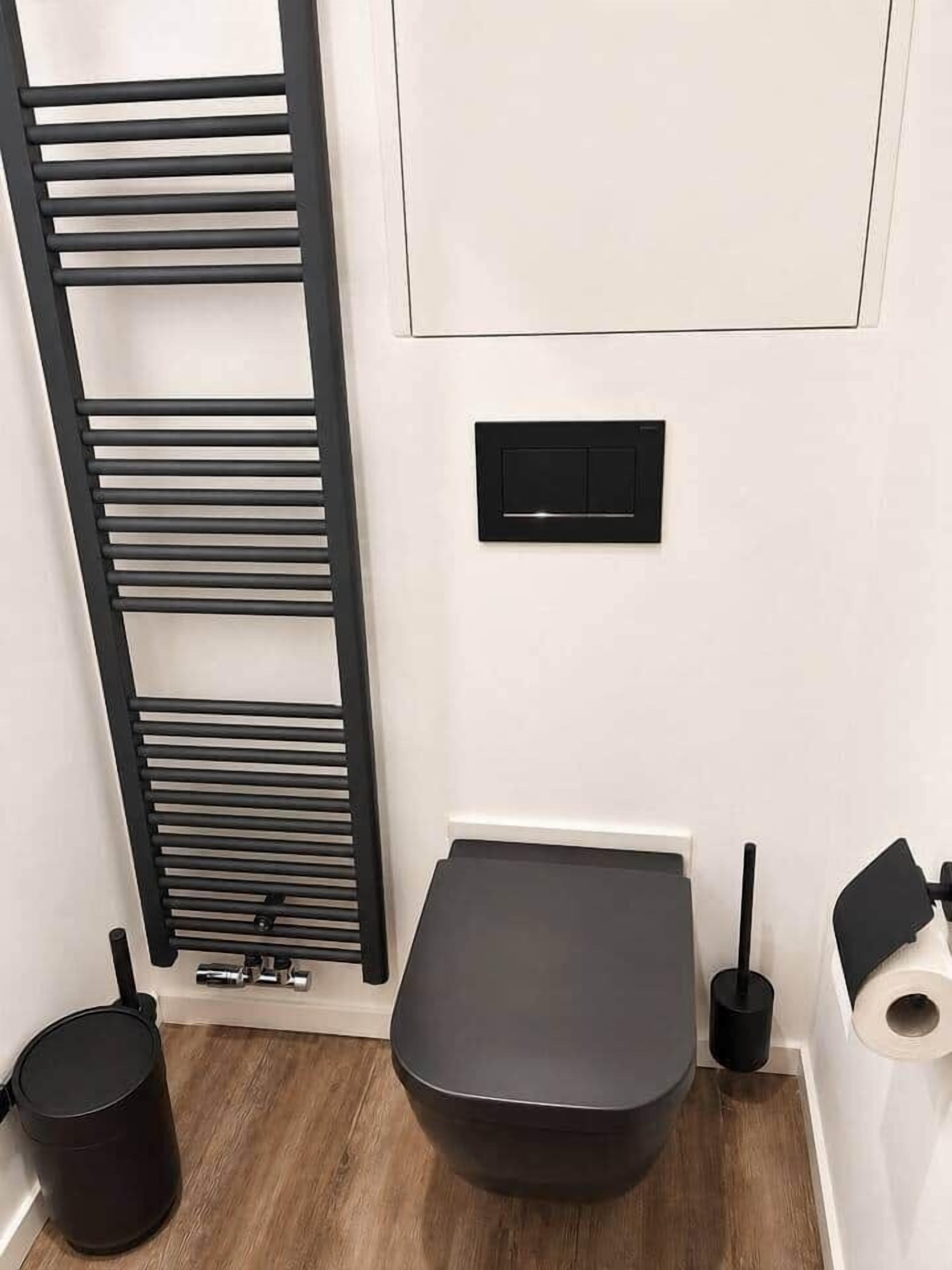 Un baño con un inodoro, un radiador negro, un cubo de basura negro y un dispensador de papel higiénico.