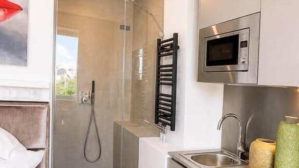 A compact hotel room with a shower, a bed, a kitchenette with a microwave, and a small window. - The Faces | Compact Studio | 305 (Berlin)