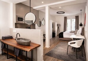 A modern hotel room with a wooden dining table, a stone basin, a bed with a white headboard, and a view of the city. - The Faces | Premium Studio | Balcony | 302 (Berlin)